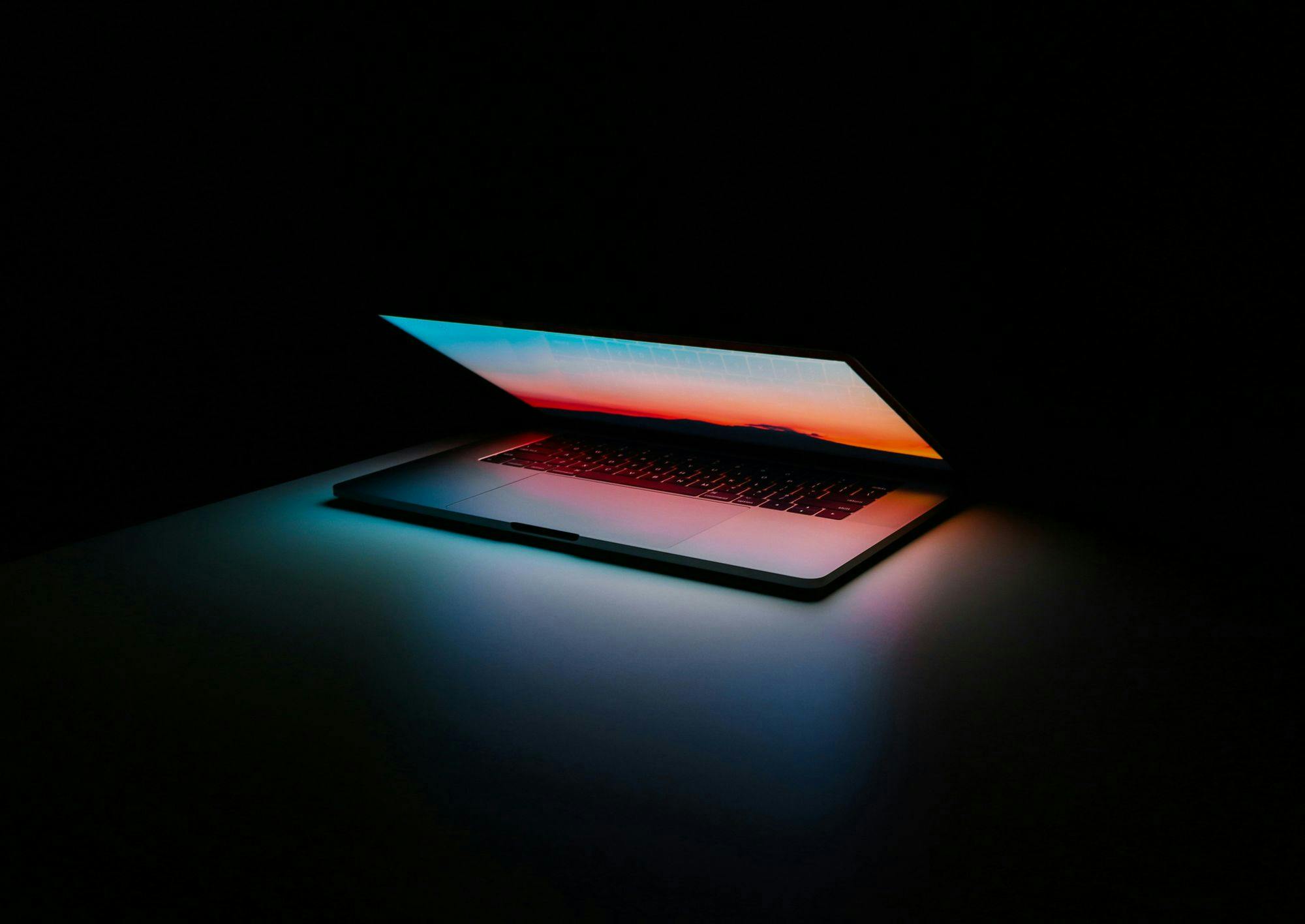 A modern laptop with a colorful backlit keyboard, emitting neon blue and orange hues in a dark environment, giving it a futuristic and sleek appearance.
